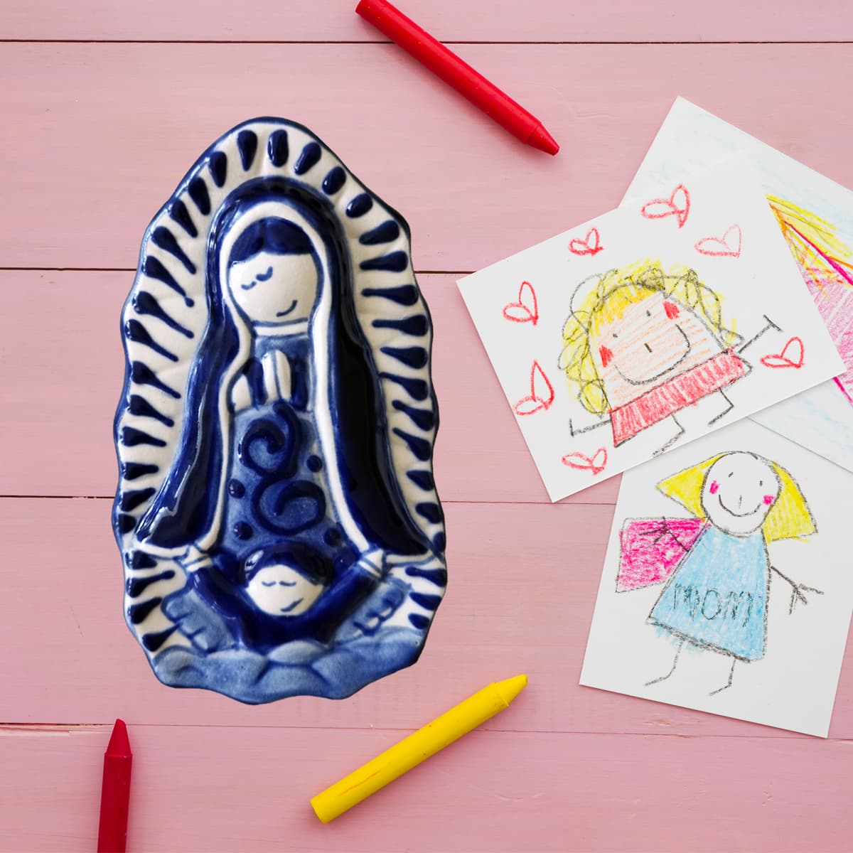 A Mexican Talavera pottery miniature of the Virgin of Guadalupe, displayed alongside colorful pencils and papers on a pink background.