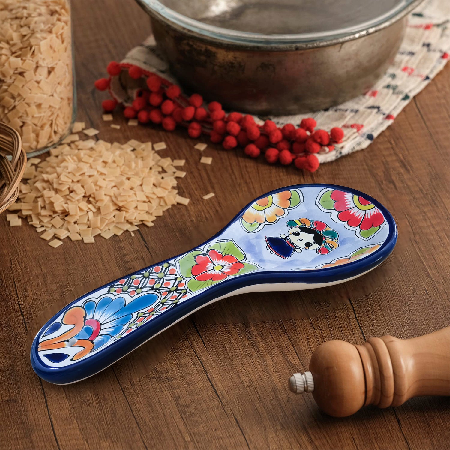 A ceramic spoon rest with a blue border and a colorful floral design, hand-painted Lele doll Talavera style, placed on a wooden surface with a background of rice and spices.
