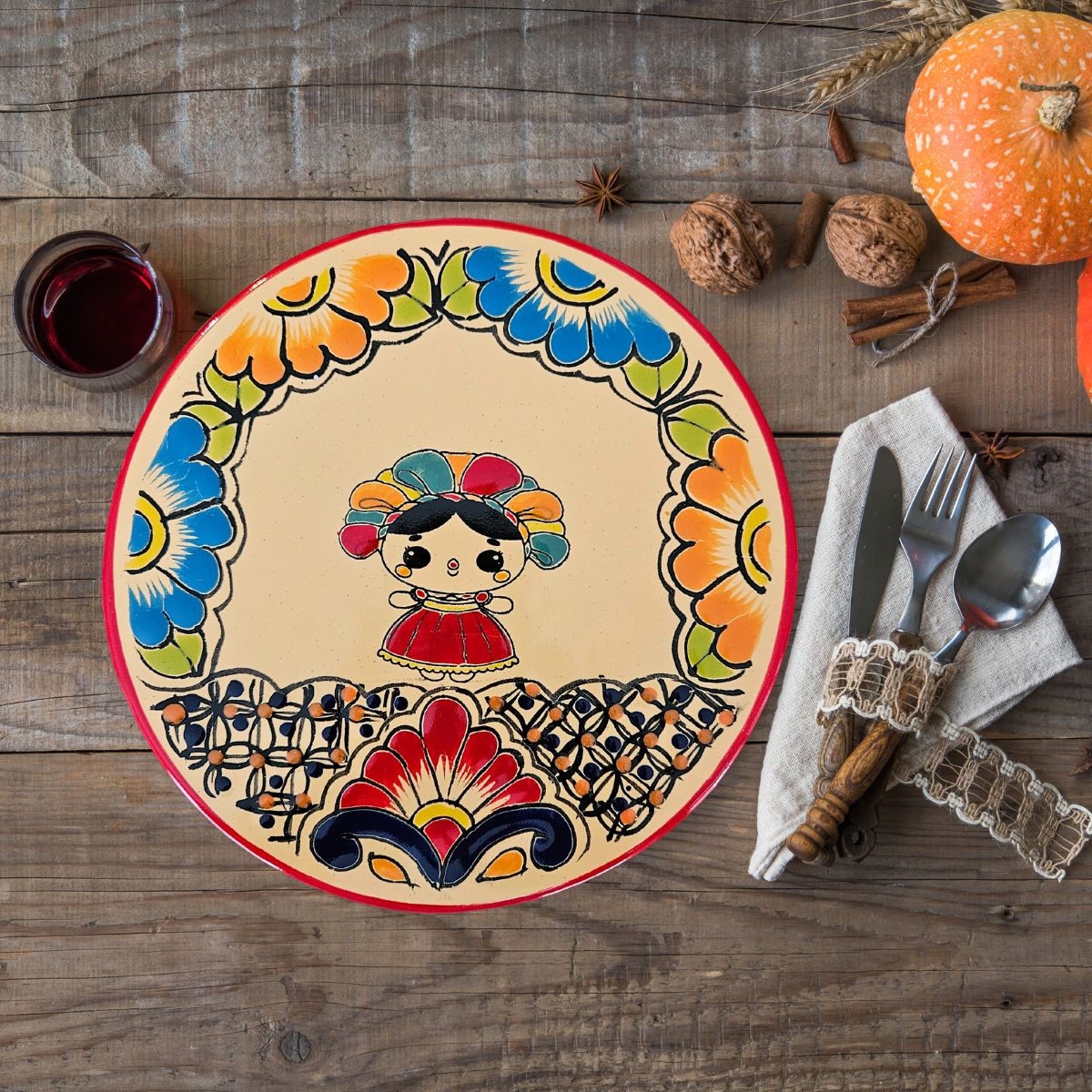 A round ceramic trivet with a floral and Lele doll patterns design, placed on a wooden surface with a fork, knife, and pumpkin decorations around it.
